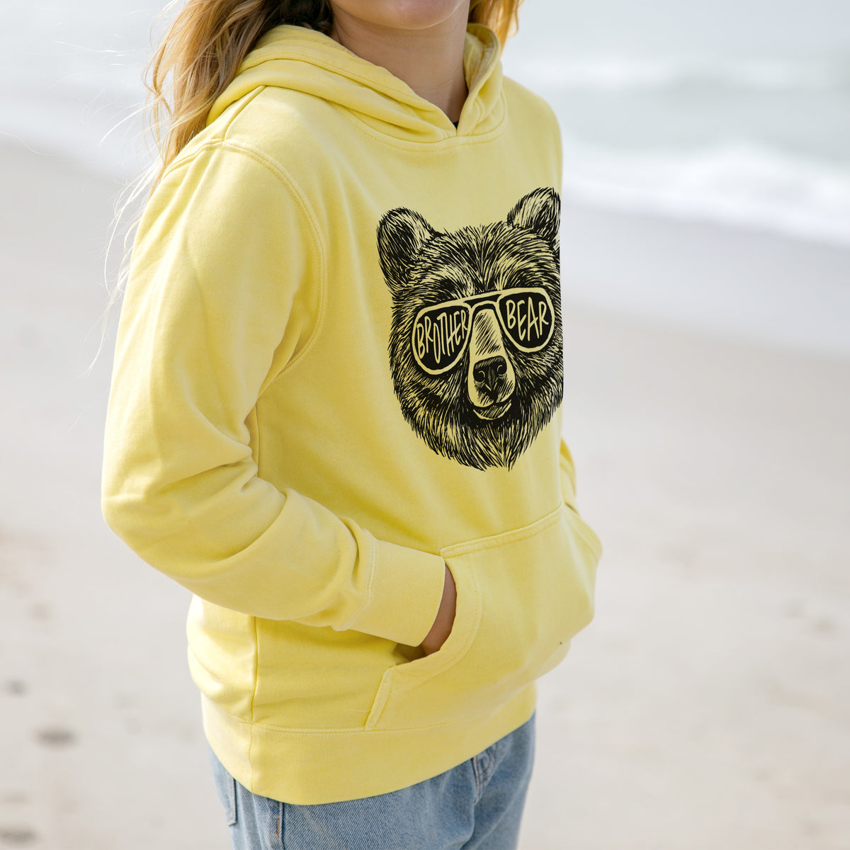 Girl wearing a youth pigment dyed hoodie printed with a black ink bear's face wearing sunglasses that read Brother Bear.