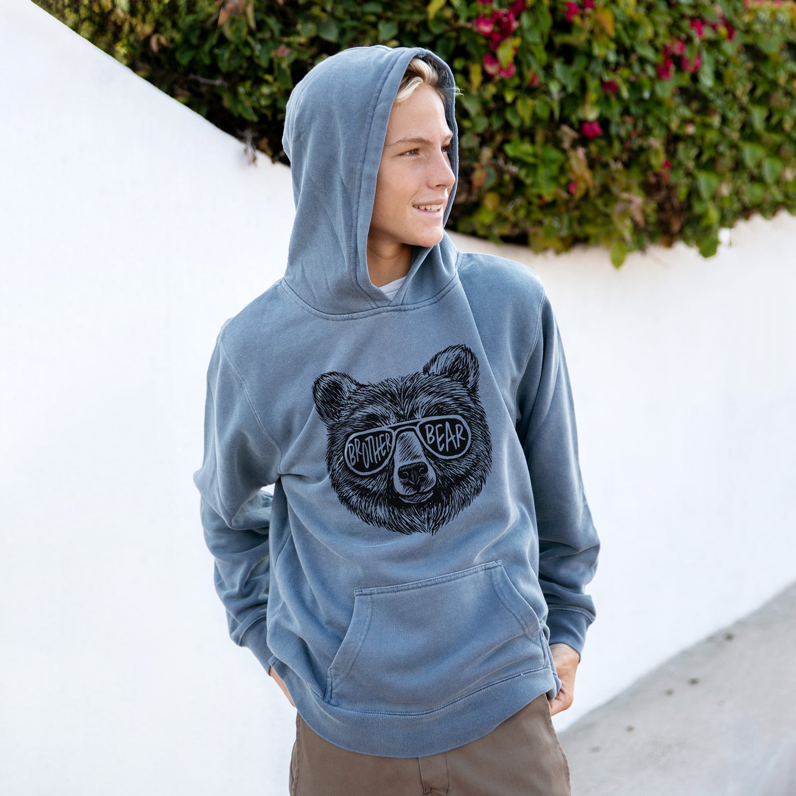 Girl wearing a youth pigment dyed hoodie printed with a black ink bear's face wearing sunglasses that read Brother Bear.