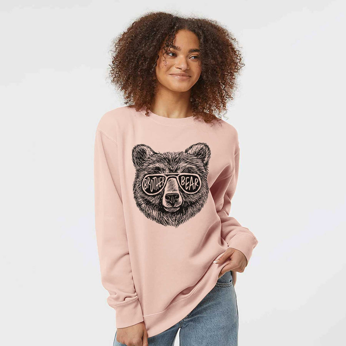 Model wearing a dusty pink pigment dyed crewneck sweatshirt printed with a black ink bear's face wearing sunglasses that read Brother Bear.