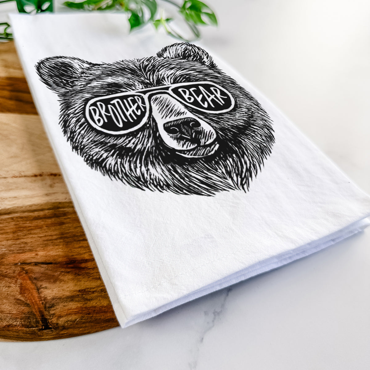 Close-up of a folded white tea towel on a wooden board, showing a black ink bear's face wearing sunglasses that read Brother Bear.