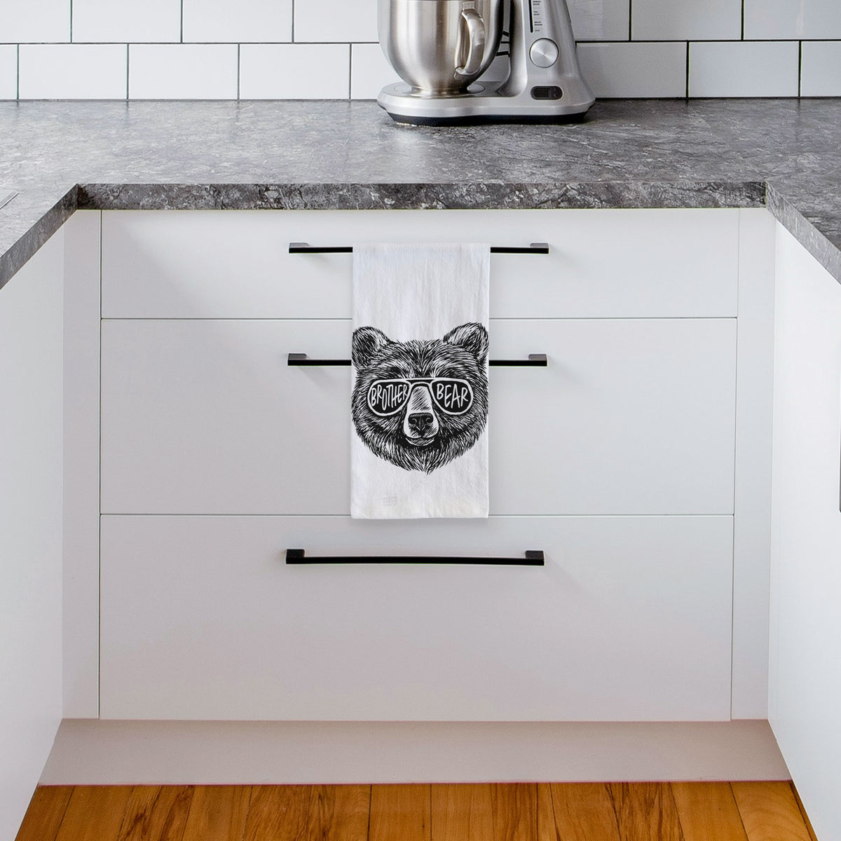 White tea towel printed with a black ink bear's face wearing sunglasses that read Brother Bear, hanging from a drawer handle in a bright white kitchen.