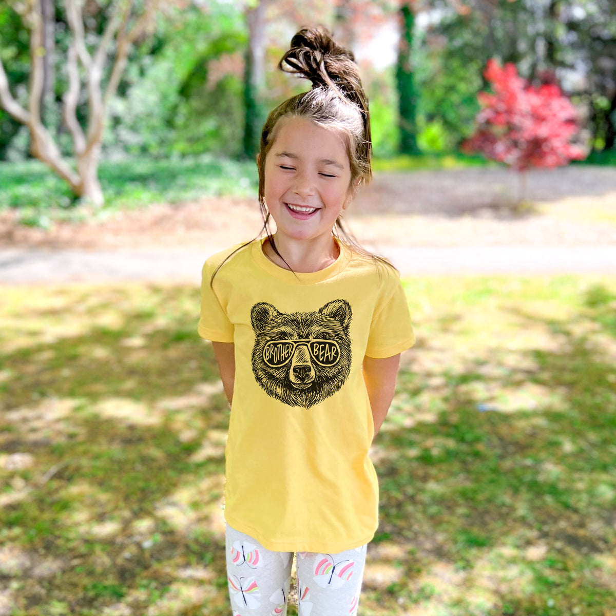 Smiling girl wearing a youth t-shirt printed with a black ink bear's face wearing sunglasses that read Brother Bear, outdoors.