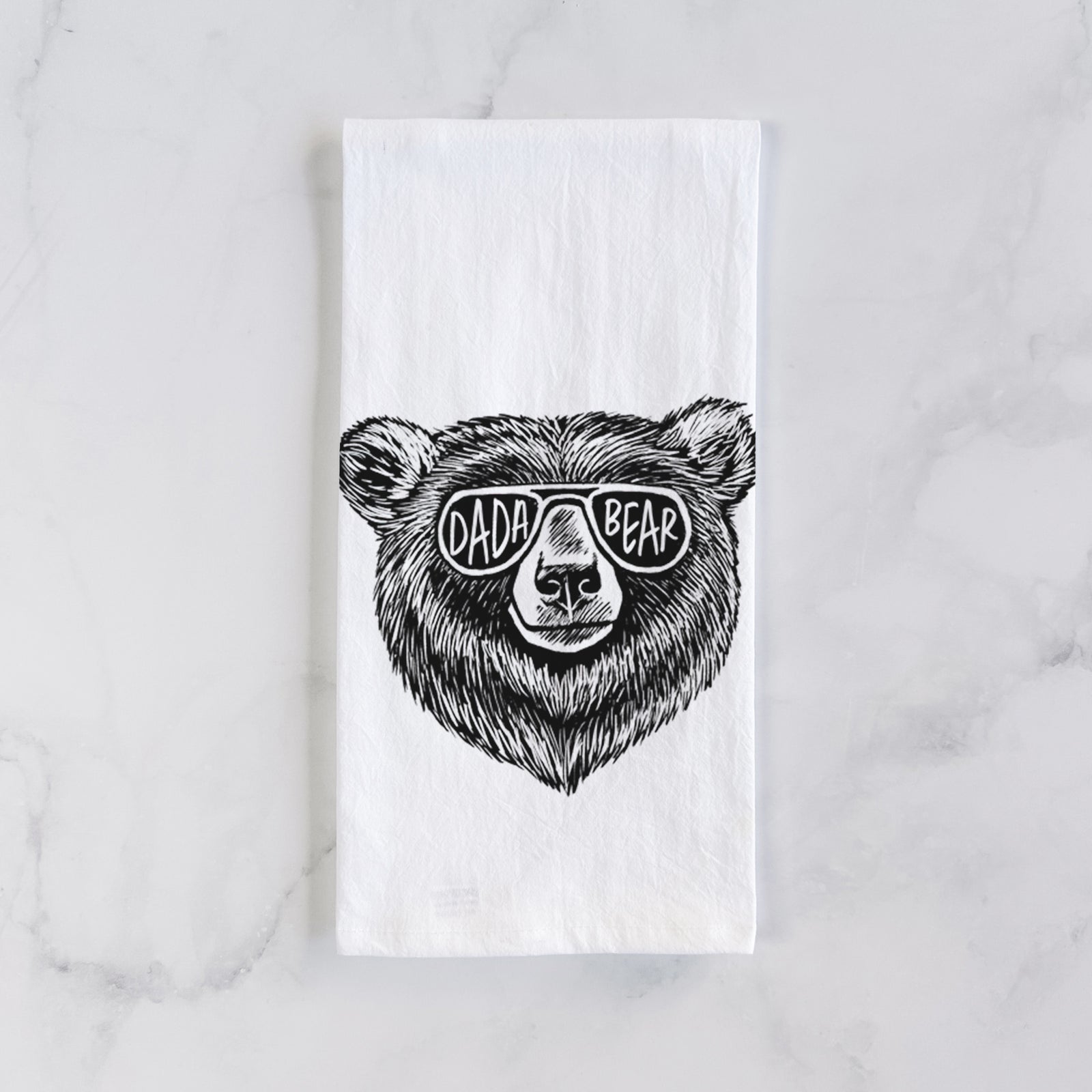 White cotton tea towel printed with a black ink bear's face wearing sunglasses that read Dada Bear, on a marble counter beside a round wooden cutting board and greenery.