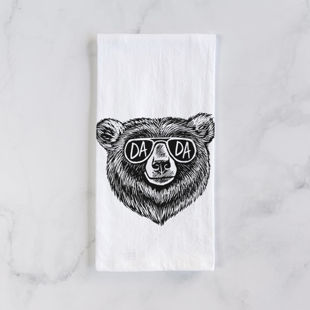 White cotton tea towel laid flat on marble, printed with a black ink bear's face wearing sunglasses that read Dada.