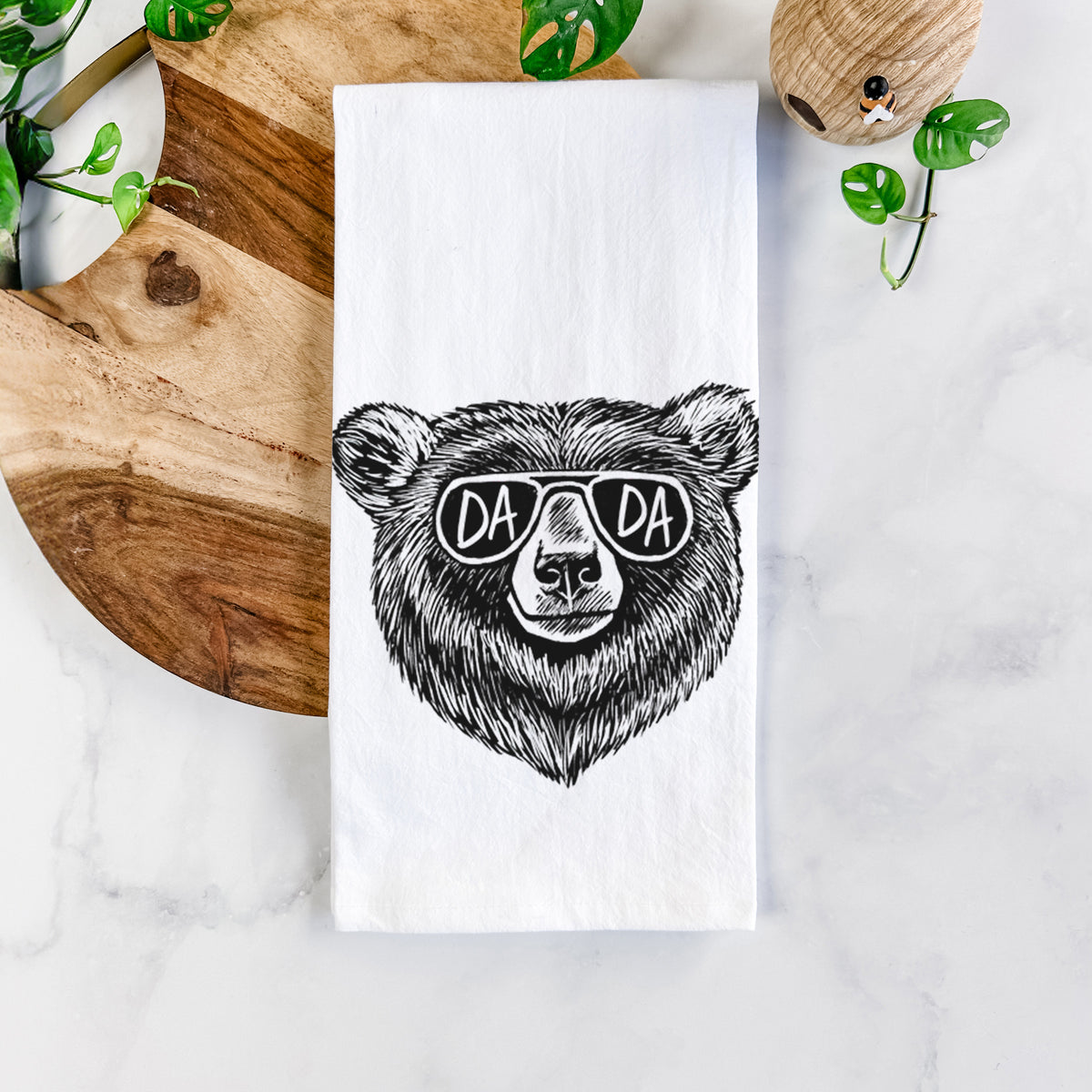 White cotton tea towel printed with a black ink bear's face wearing sunglasses that read Dada, on a marble counter beside a round wooden cutting board and greenery.