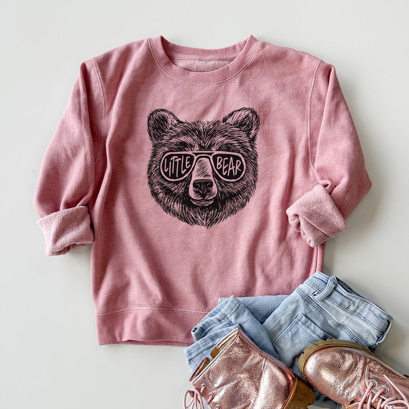 Boy wearing a youth crewneck sweatshirt printed with a black ink bear's face wearing sunglasses that read Little Bear, outdoors.