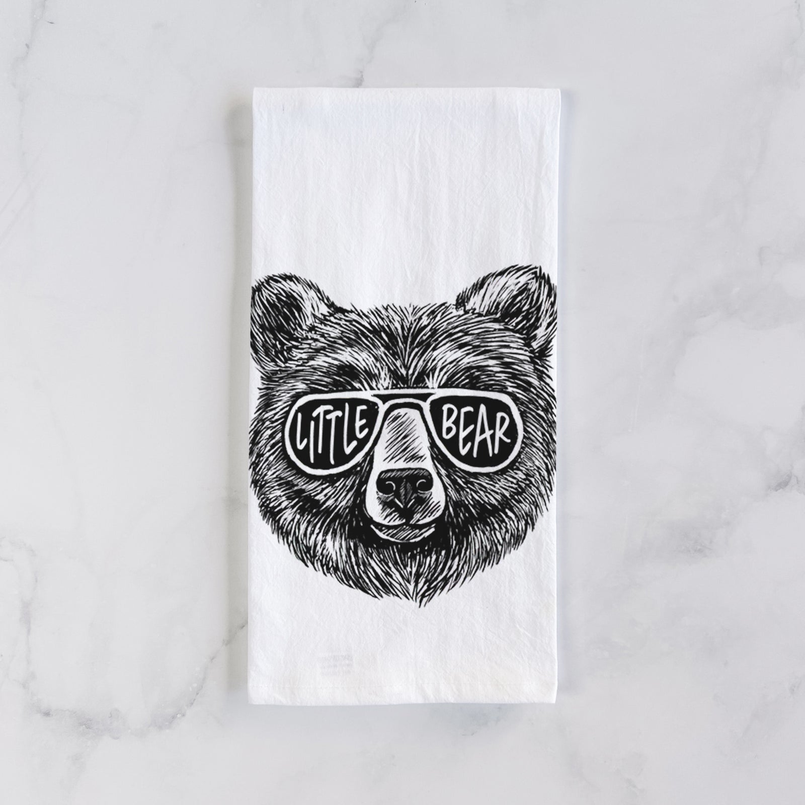 White cotton tea towel printed with a black ink bear's face wearing sunglasses that read Little Bear, on a marble counter beside a round wooden cutting board and greenery.