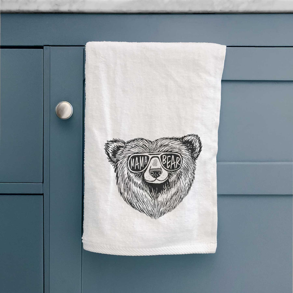 White hand towel printed with a black ink bear's face wearing sunglasses that read Mama Bear, hanging on a cabinet door.