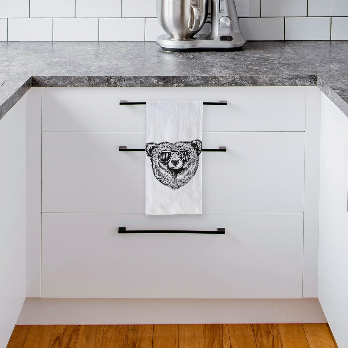 White tea towel printed with a black ink bear's face wearing sunglasses that read Mama Bear, hanging from a drawer handle in a bright white kitchen.