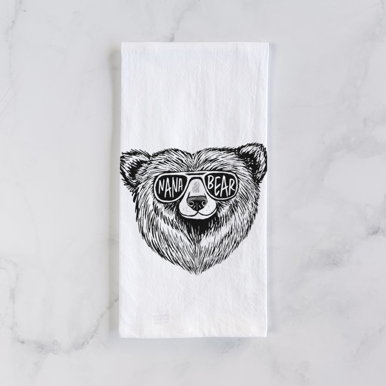 White cotton tea towel printed with a black ink bear's face wearing sunglasses that read Nana Bear, on a marble counter beside a round wooden cutting board and greenery.