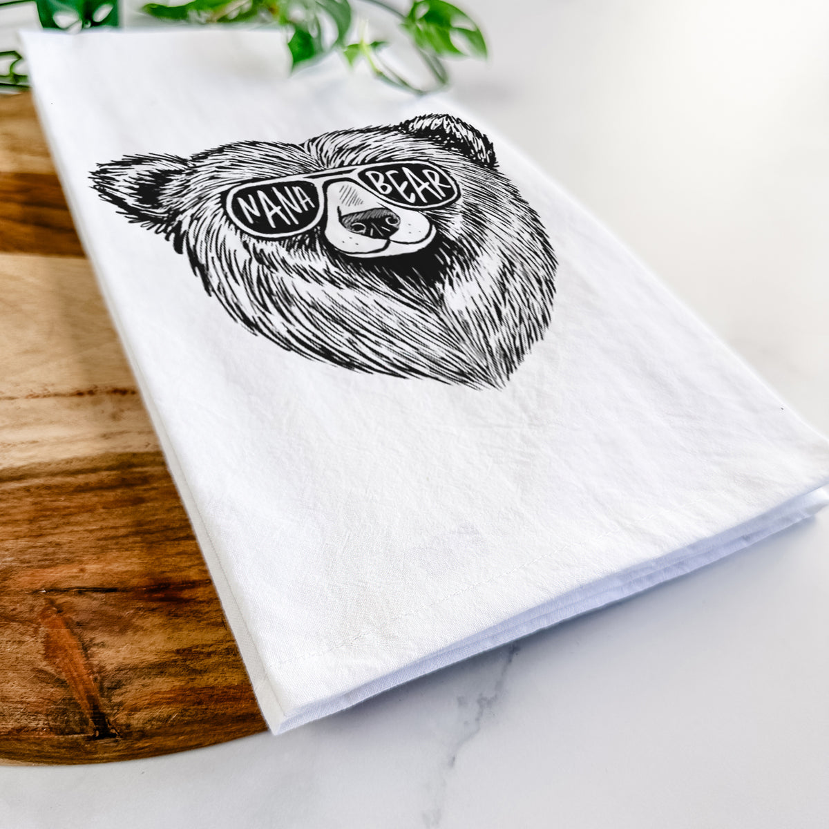 Close-up of a folded white tea towel on a wooden board, showing a black ink bear's face wearing sunglasses that read Nana Bear.