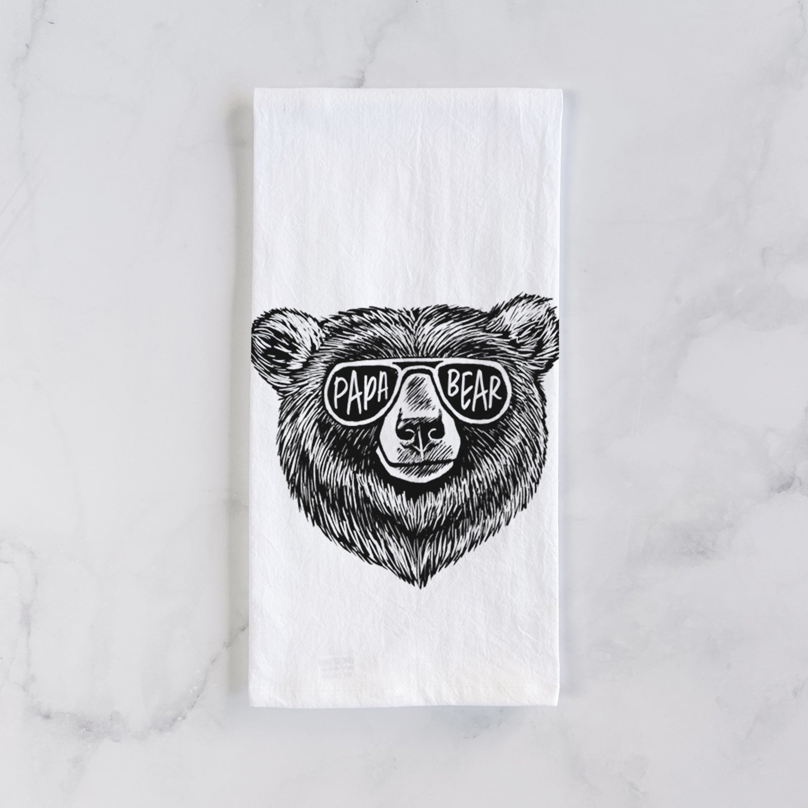 White cotton tea towel printed with a black ink bear's face wearing sunglasses that read Papa Bear, on a marble counter beside a round wooden cutting board and greenery.