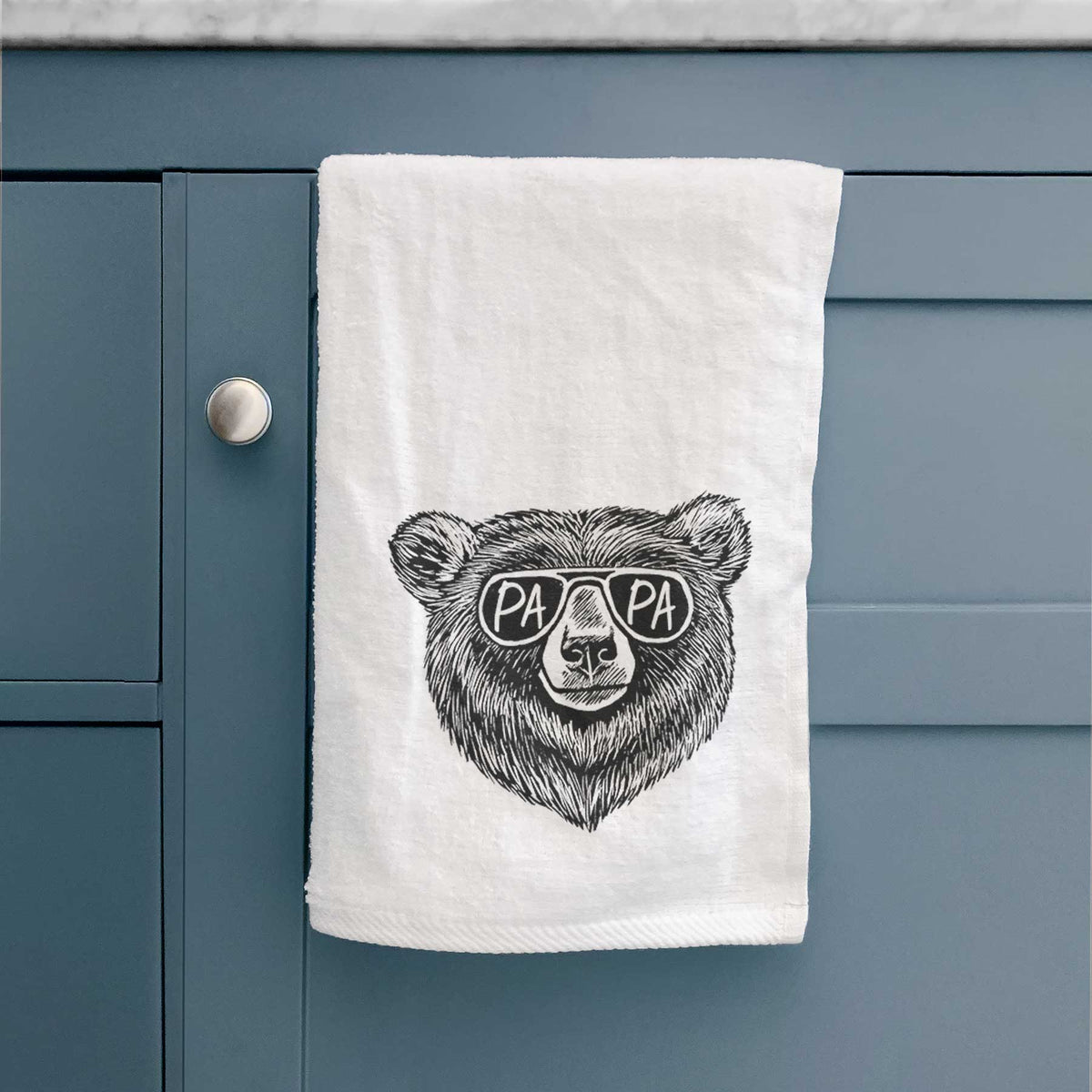 White hand towel printed with a black ink bear's face wearing sunglasses that read Papa, hanging on a cabinet door.