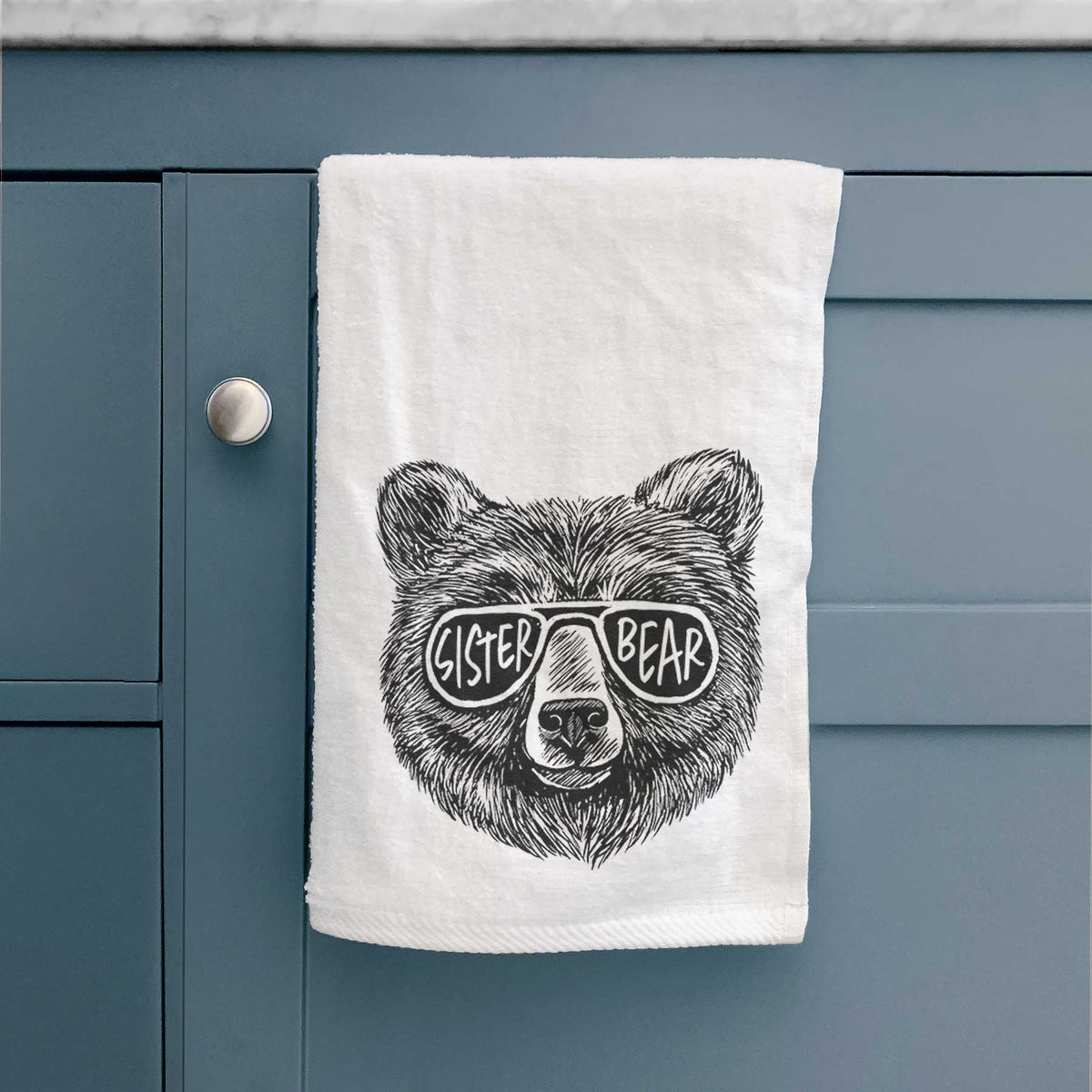 White hand towel printed with a black ink bear's face wearing sunglasses that read Sister Bear, hanging on a cabinet door.