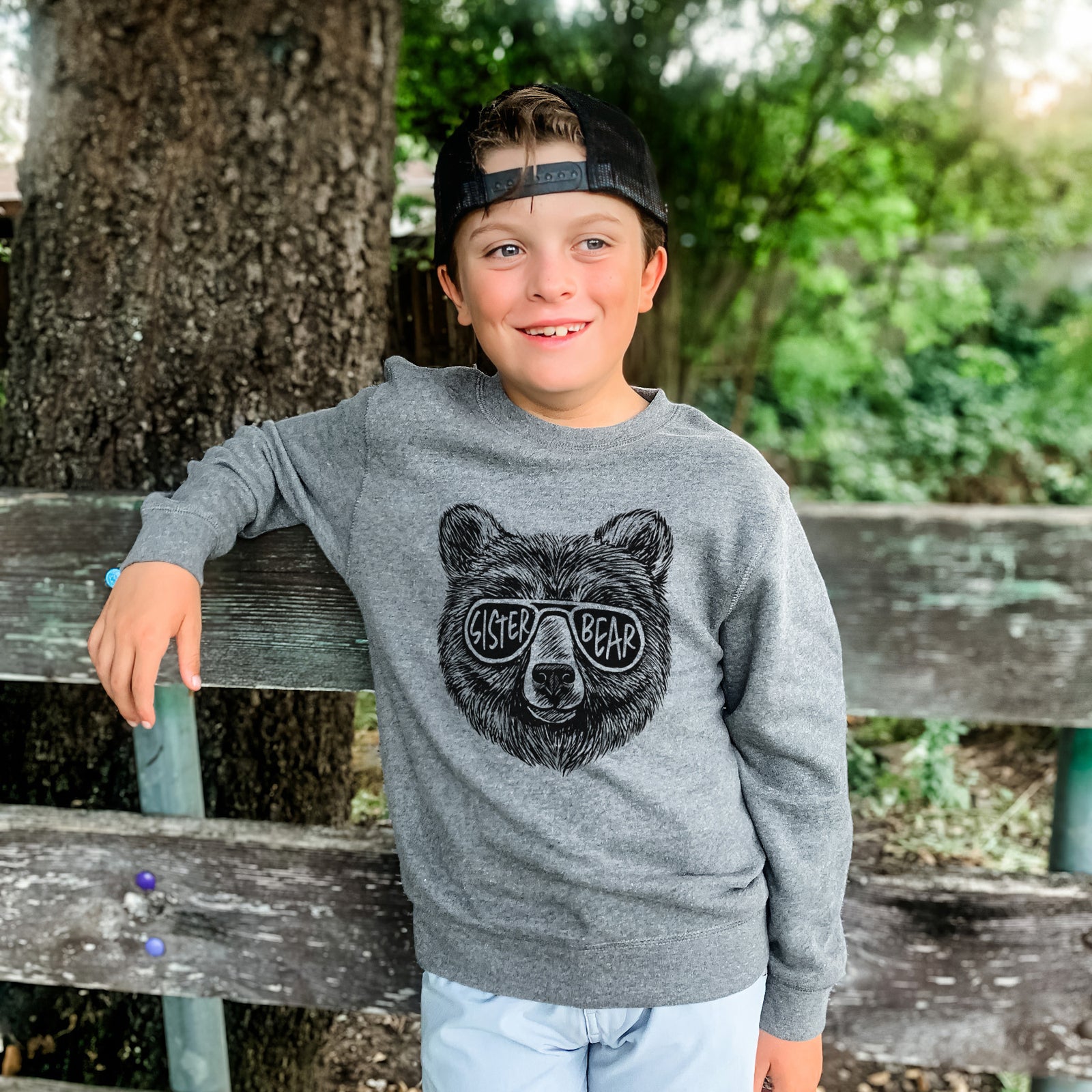 Boy wearing a youth crewneck sweatshirt printed with a black ink bear's face wearing sunglasses that read Sister Bear, outdoors.