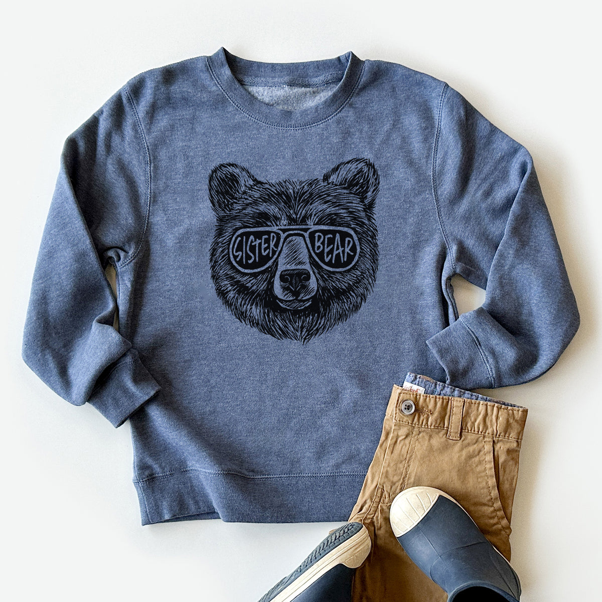 Youth crewneck sweatshirt printed with a black ink bear's face wearing sunglasses that read Sister Bear, styled flat with khaki pants and shoes.