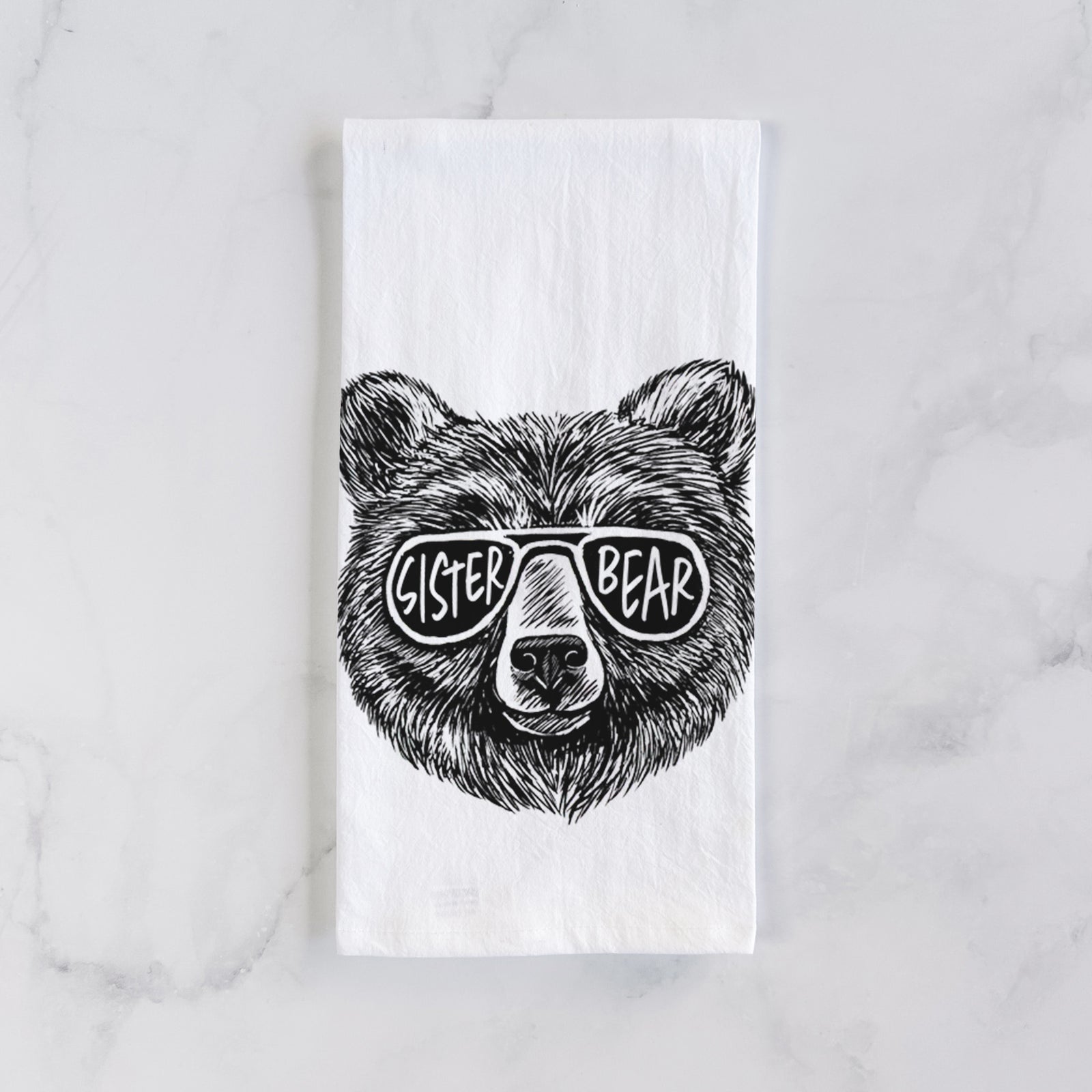 White cotton tea towel printed with a black ink bear's face wearing sunglasses that read Sister Bear, on a marble counter beside a round wooden cutting board and greenery.