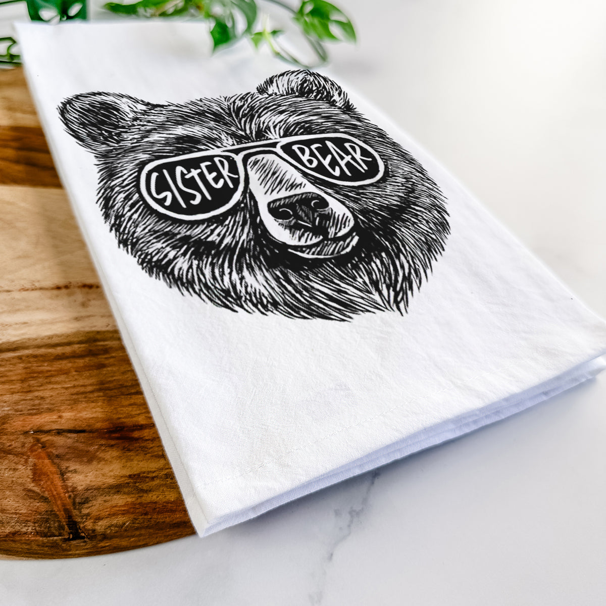 Close-up of a folded white tea towel on a wooden board, showing a black ink bear's face wearing sunglasses that read Sister Bear.
