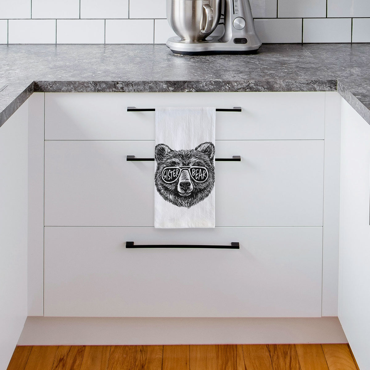 White tea towel printed with a black ink bear's face wearing sunglasses that read Sister Bear, hanging from a drawer handle in a bright white kitchen.