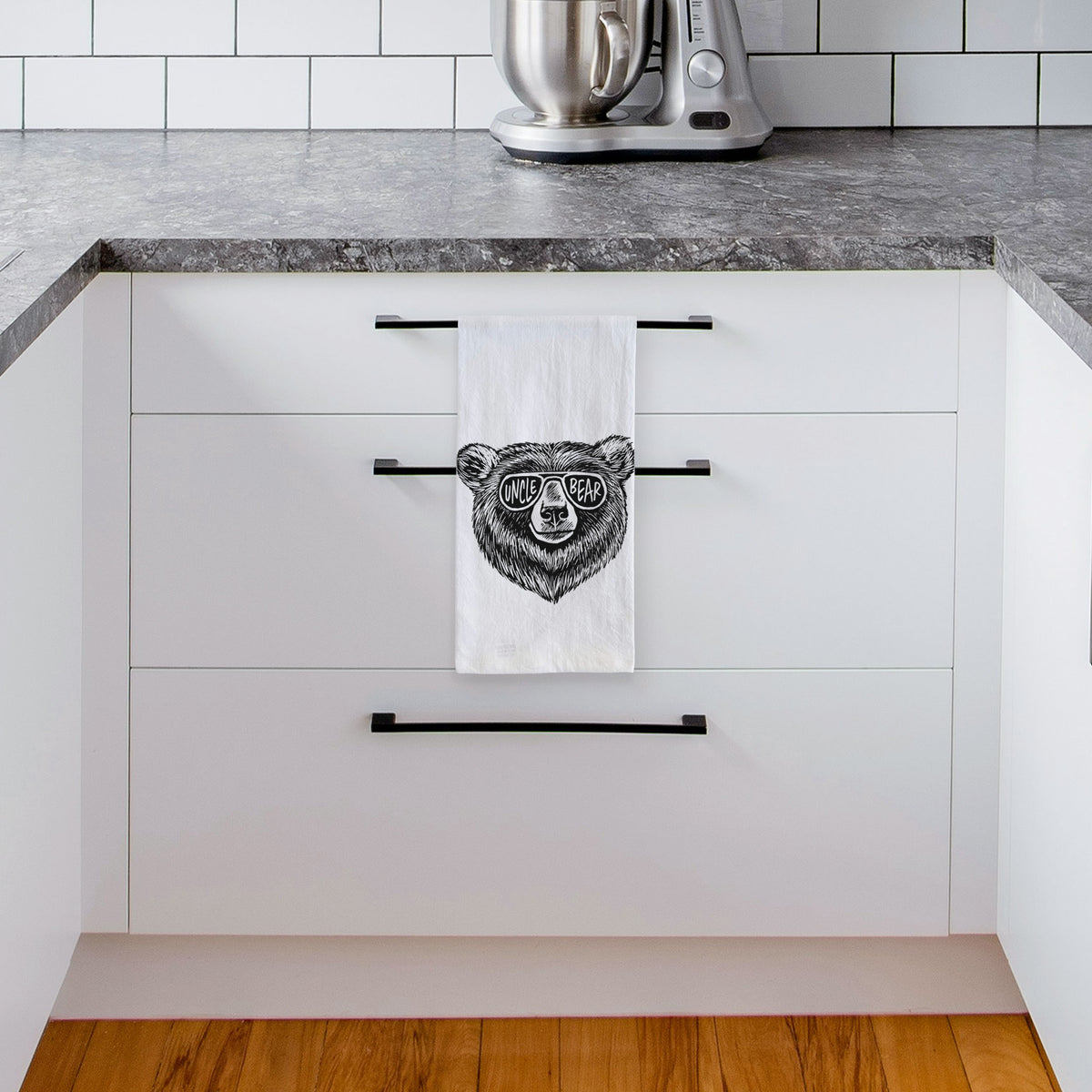White tea towel printed with a black ink bear's face wearing sunglasses that read Uncle Bear, hanging from a drawer handle in a bright white kitchen.