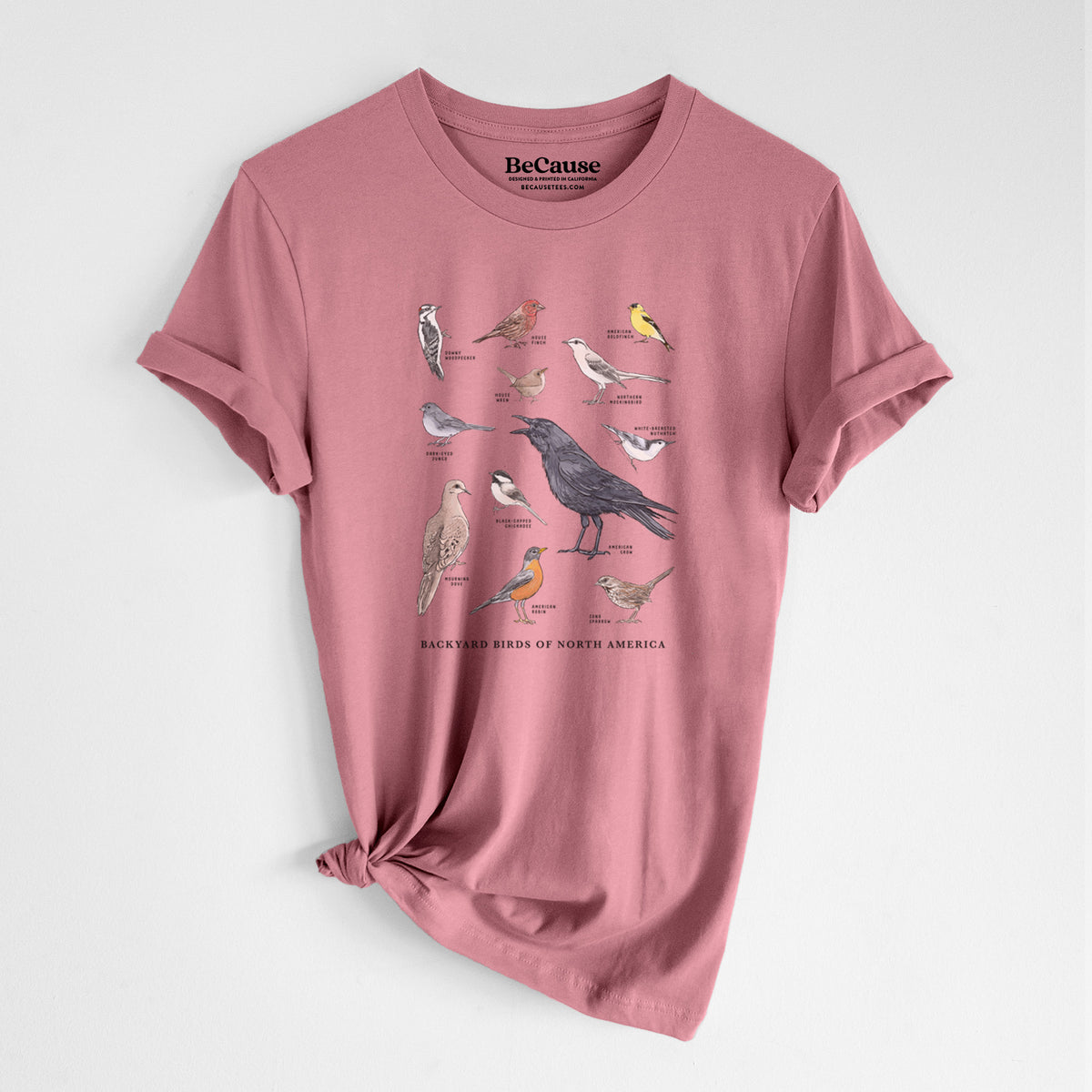 Mauve pink lightweight cotton t-shirt laid flat with the sleeves rolled, printed with a full-color chart of North American backyard birds, each labeled with its species.