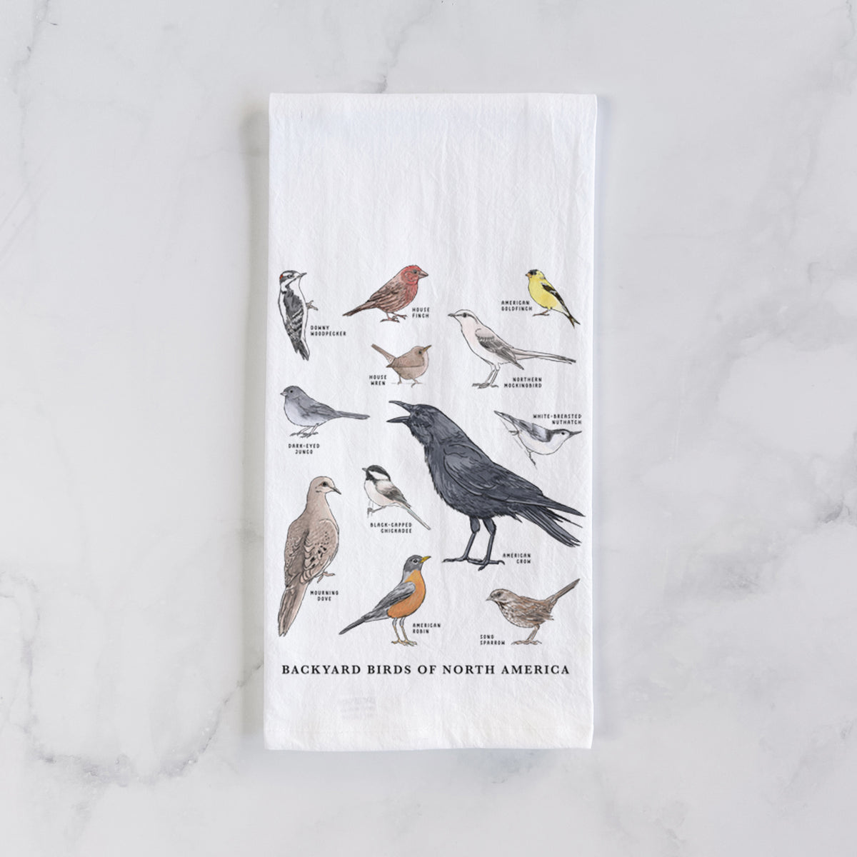 White cotton tea towel laid flat on marble, printed with a full-color chart of North American backyard birds, each labeled with its species.