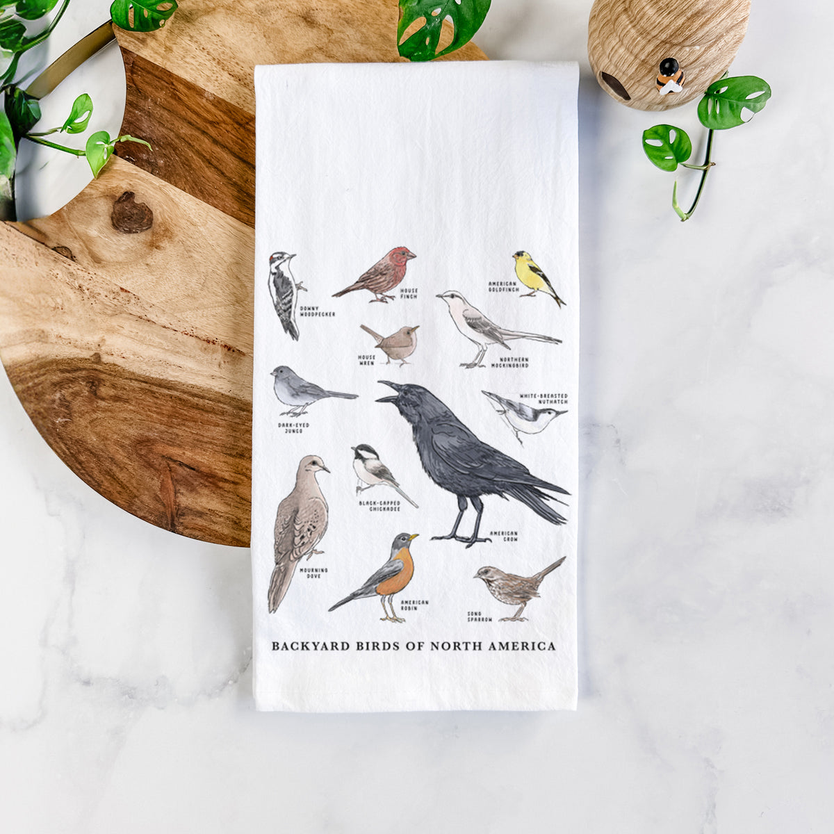 White cotton tea towel printed with a full-color chart of North American backyard birds, each labeled with its species, on a marble counter beside a round wooden cutting board and greenery.