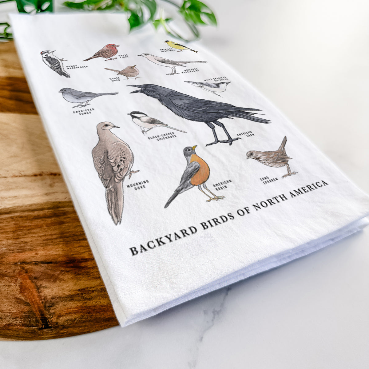 Close-up of a folded white tea towel on a wooden board, showing a full-color chart of North American backyard birds, each labeled with its species.