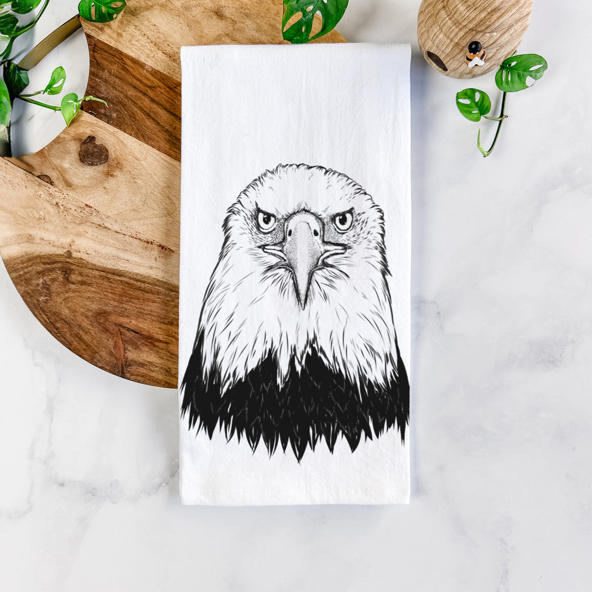 White cotton tea towel printed with a detailed black ink portrait of a bald eagle facing forward, on a marble counter beside a round wooden cutting board and greenery.
