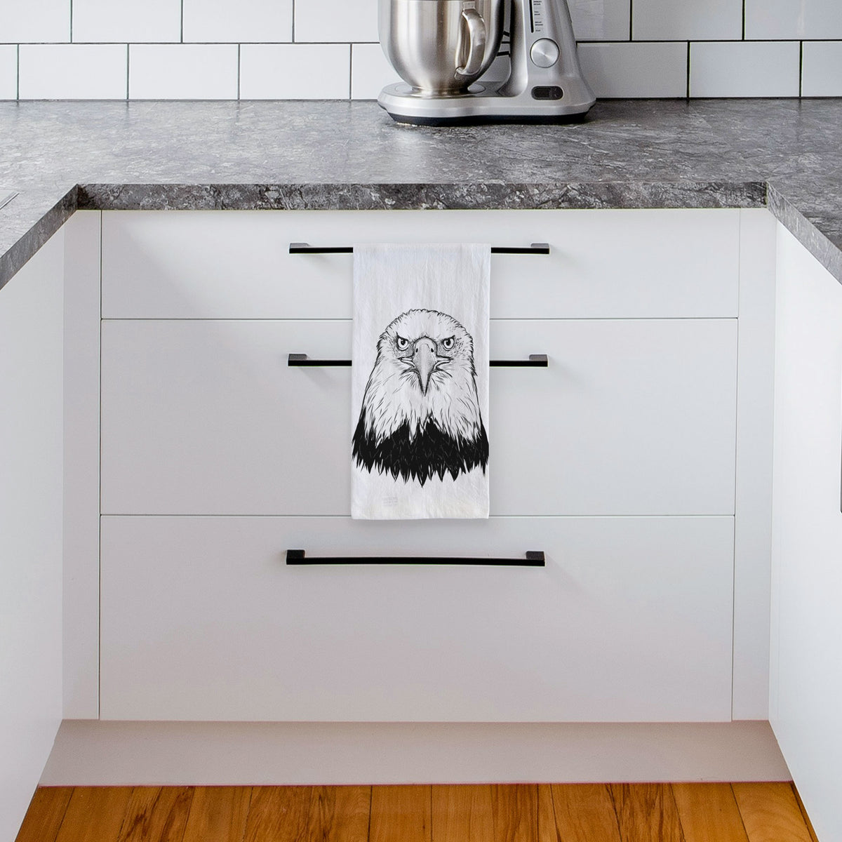 White tea towel printed with a detailed black ink portrait of a bald eagle facing forward, hanging from a drawer handle in a bright white kitchen.