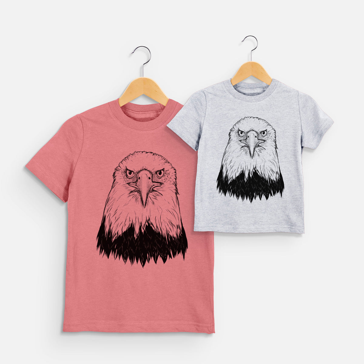 Two youth t-shirts in different colors on hangers, printed with a detailed black ink portrait of a bald eagle facing forward.