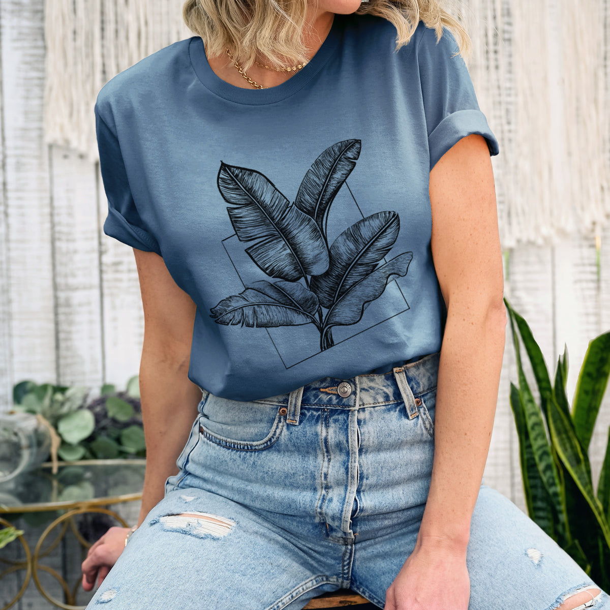 Woman wearing a steel blue lightweight cotton t-shirt printed with a black ink illustration of banana leaves in front of a square outline.