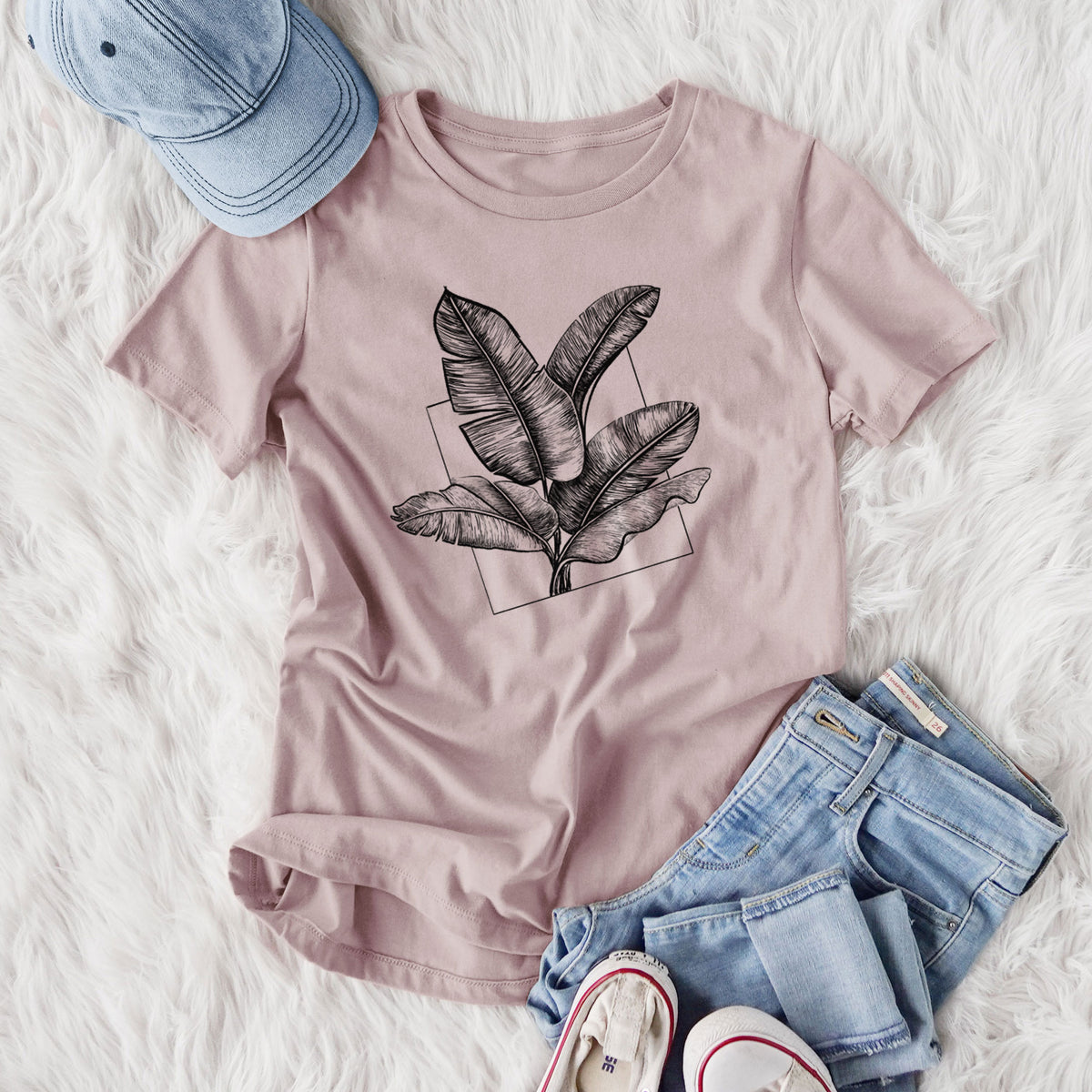 Women's relaxed fit cotton t-shirt printed with a black ink illustration of banana leaves in front of a square outline, styled flat on a white faux fur rug with jeans.