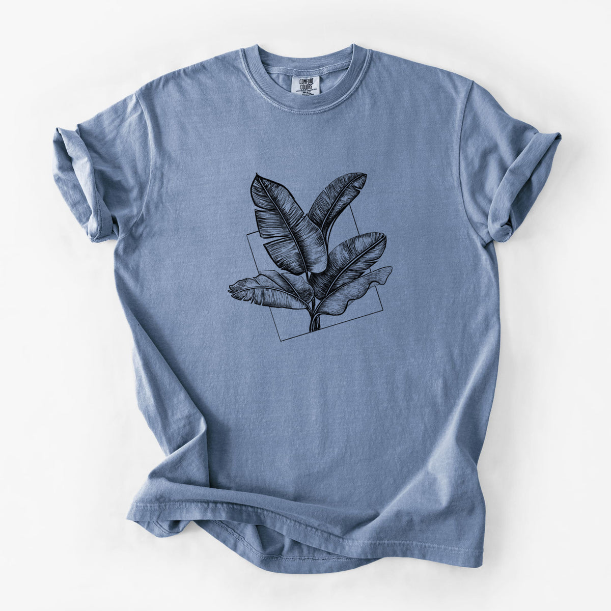 Blue jean Comfort Colors t-shirt laid flat, printed with a black ink illustration of banana leaves in front of a square outline.