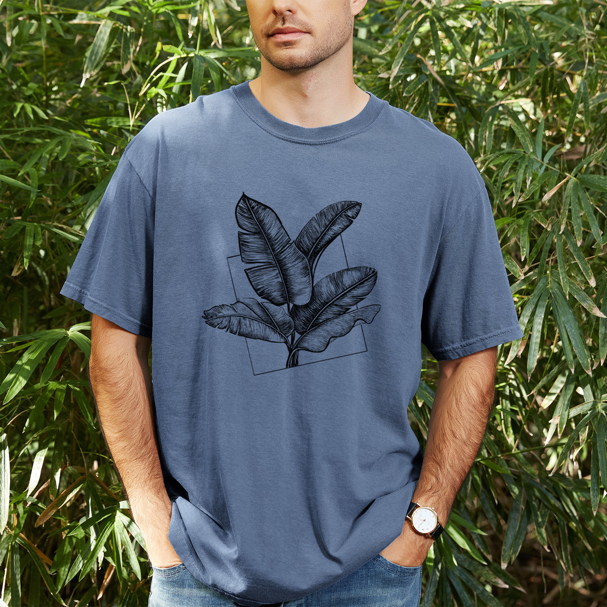 Man wearing a Comfort Colors t-shirt printed with a black ink illustration of banana leaves in front of a square outline, outdoors.