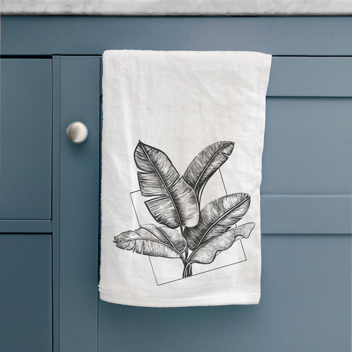 White hand towel printed with a black ink illustration of banana leaves in front of a square outline, hanging on a cabinet door.