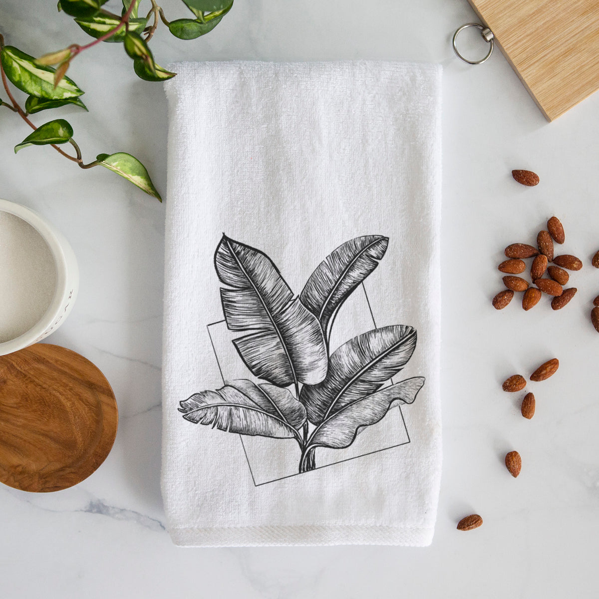White hand towel printed with a black ink illustration of banana leaves in front of a square outline.