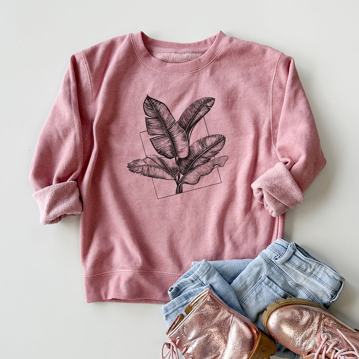 Youth crewneck sweatshirt printed with a black ink illustration of banana leaves in front of a square outline, styled flat with jeans and boots.