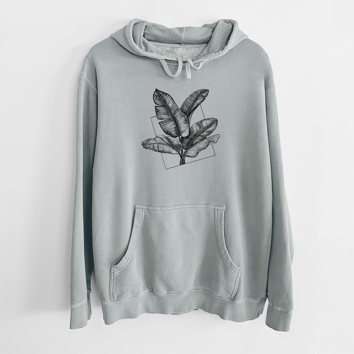 Sage green pigment dyed hoodie laid flat, printed with a black ink illustration of banana leaves in front of a square outline.