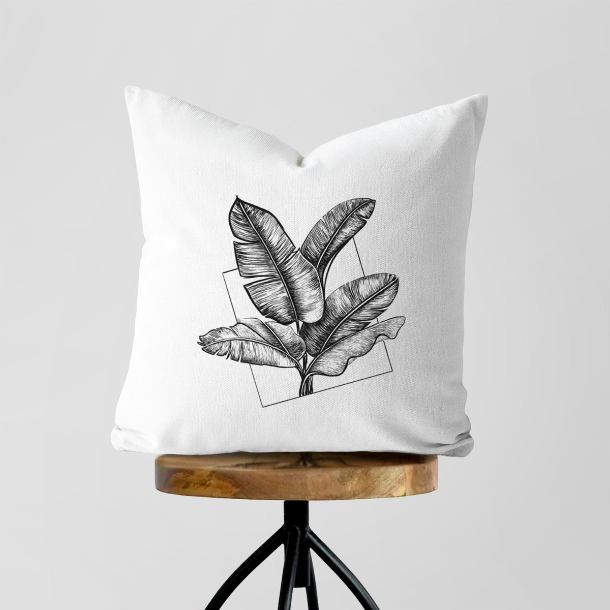 White pillow cover printed with a black ink illustration of banana leaves in front of a square outline, on a wooden stool.