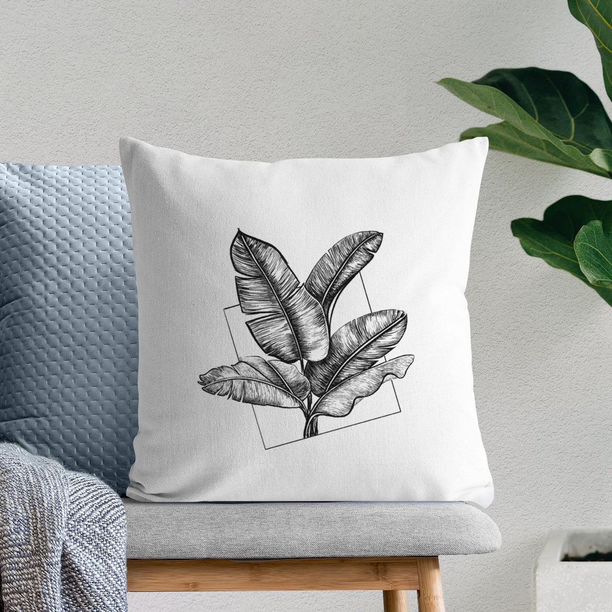 White pillow cover printed with a black ink illustration of banana leaves in front of a square outline, on a bench beside plants.