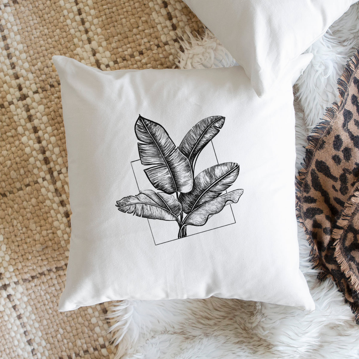 White pillow cover printed with a black ink illustration of banana leaves in front of a square outline, on a woven chair.