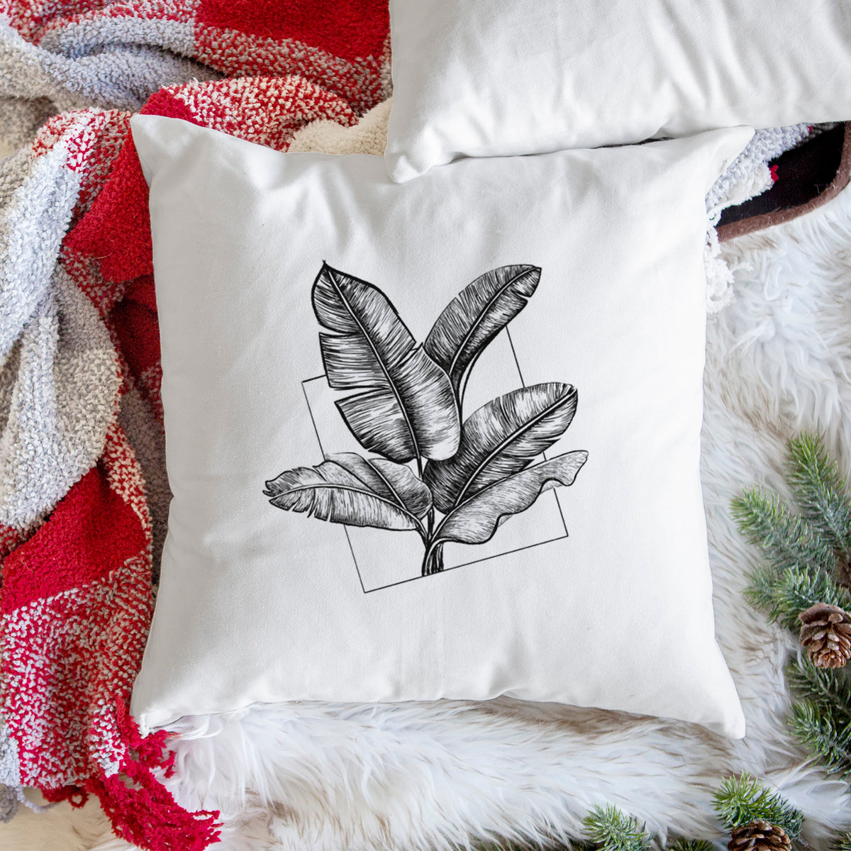 White pillow cover printed with a black ink illustration of banana leaves in front of a square outline, on a woven chair.