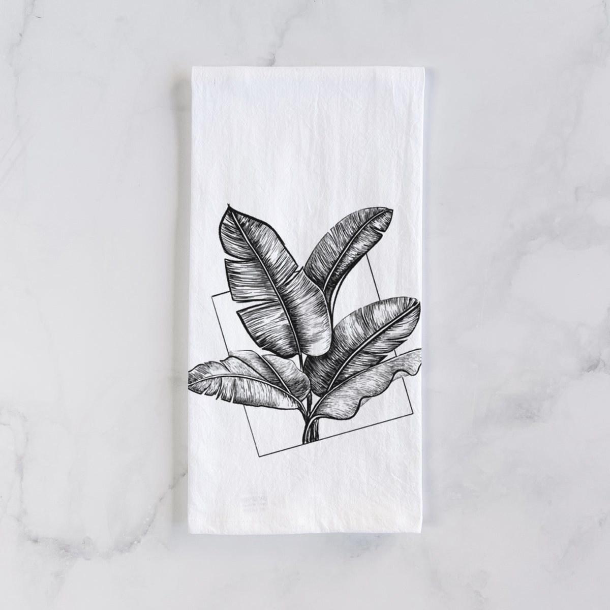 White cotton tea towel laid flat on marble, printed with a black ink illustration of banana leaves in front of a square outline.