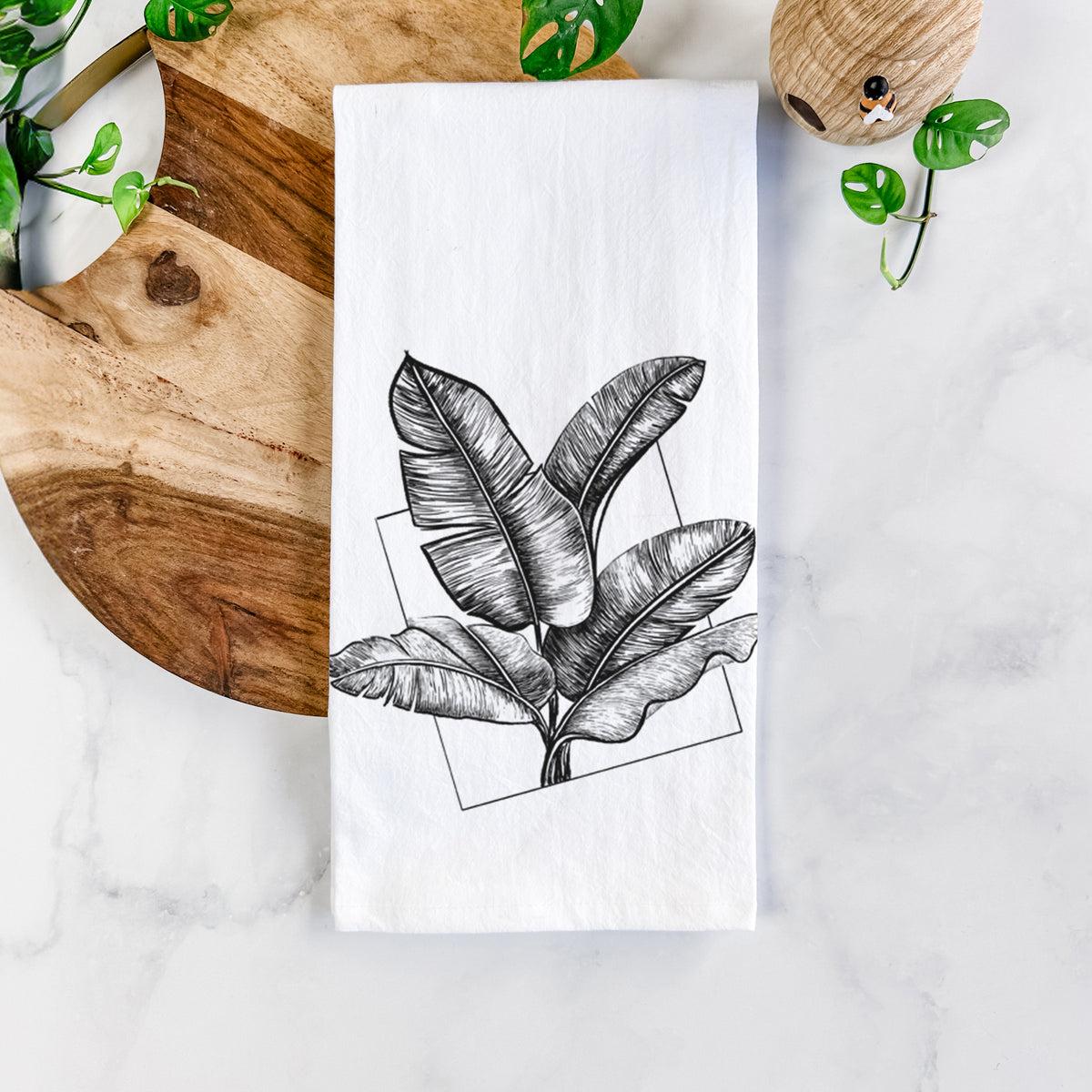 White cotton tea towel printed with a black ink illustration of banana leaves in front of a square outline, on a marble counter beside a round wooden cutting board and greenery.