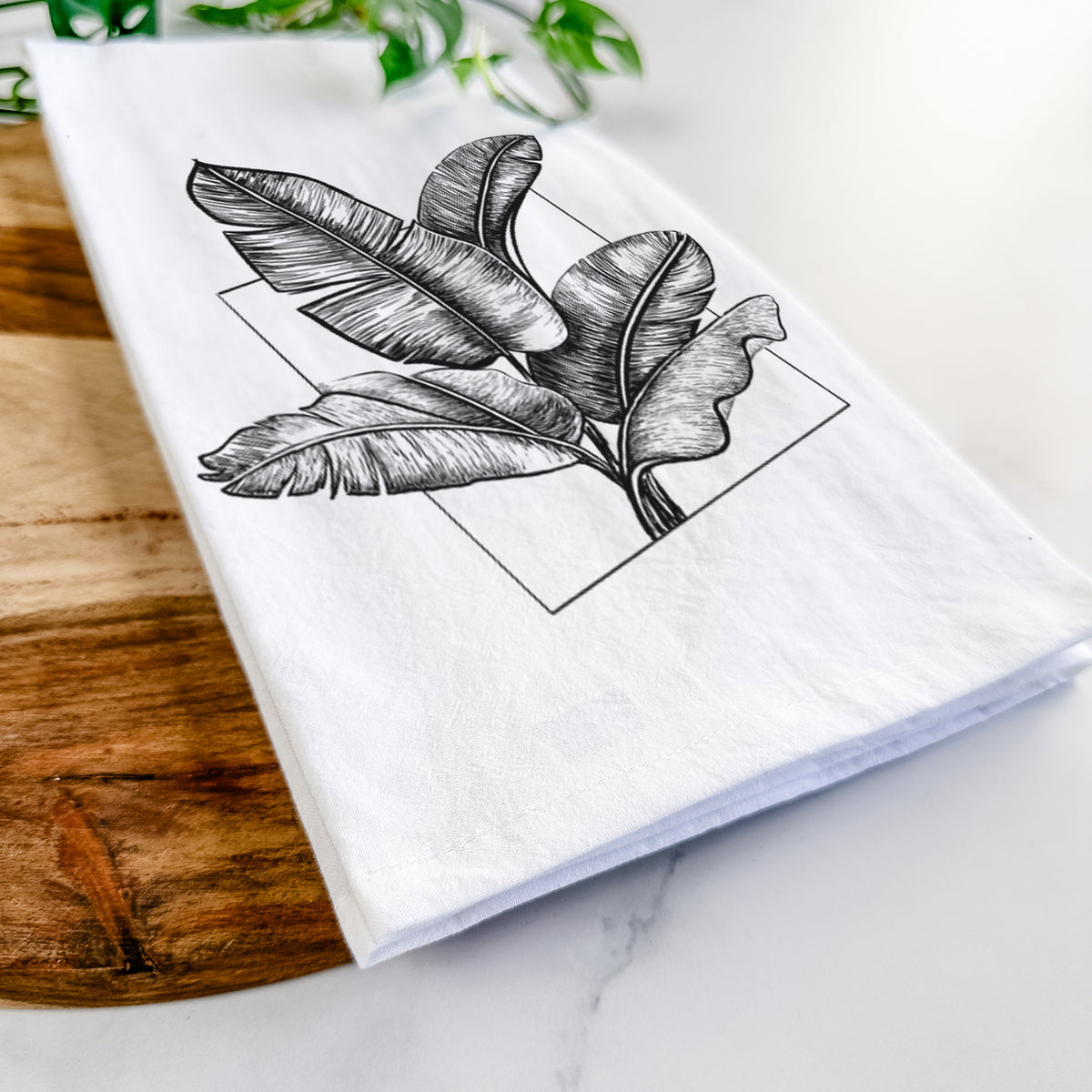 Close-up of a folded white tea towel on a wooden board, showing a black ink illustration of banana leaves in front of a square outline.