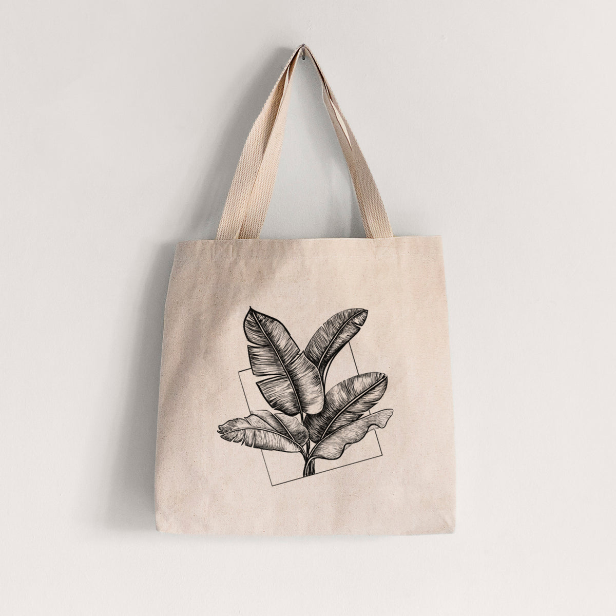 Natural canvas tote bag printed with a black ink illustration of banana leaves in front of a square outline.