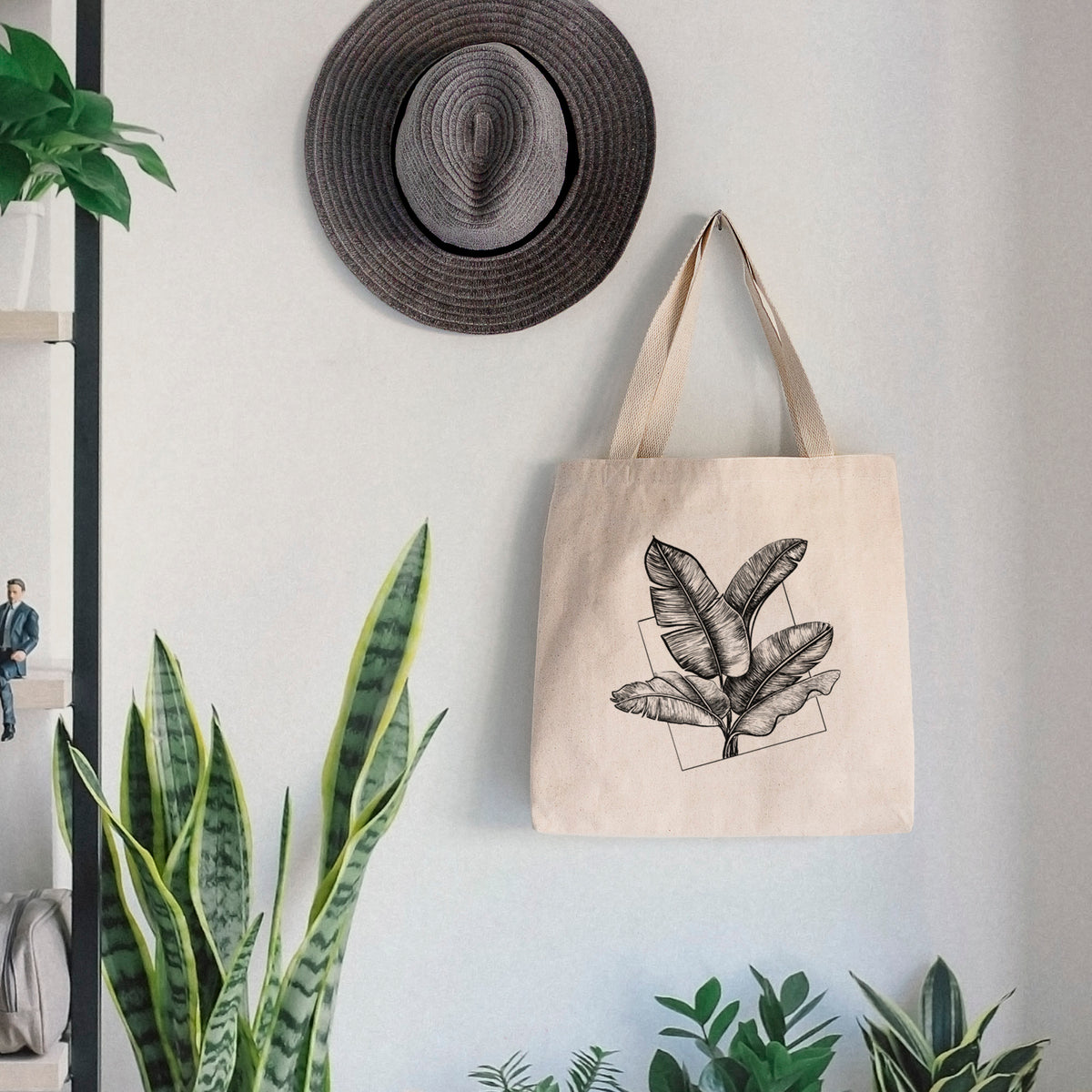 Canvas tote bag printed with a black ink illustration of banana leaves in front of a square outline, hanging on a wall hook below a hat, next to potted plants.