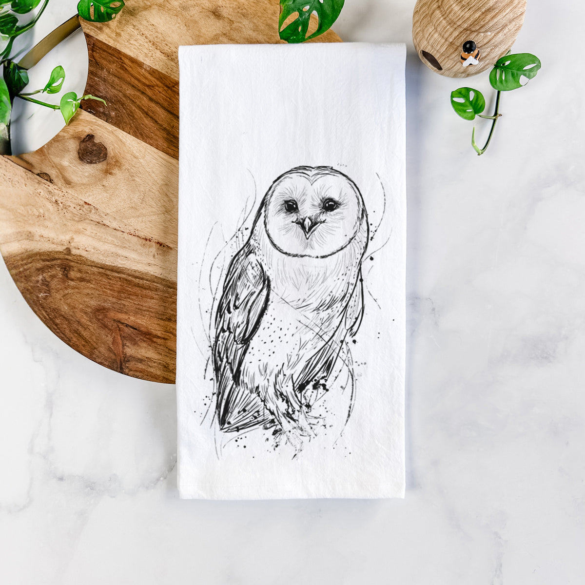 White cotton tea towel printed with a detailed black ink illustration of a barn owl standing and facing forward, on a marble counter beside a round wooden cutting board and greenery.