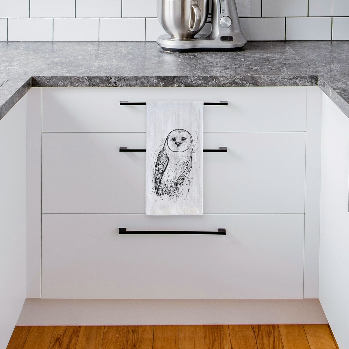White tea towel printed with a detailed black ink illustration of a barn owl standing and facing forward, hanging from a drawer handle in a bright white kitchen.